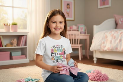 Little girl wearing Bows and Bling It’s a Cheer Thing t-shirt, holding glittery hair bow with pom poms on bedroom floor, cheer-themed room